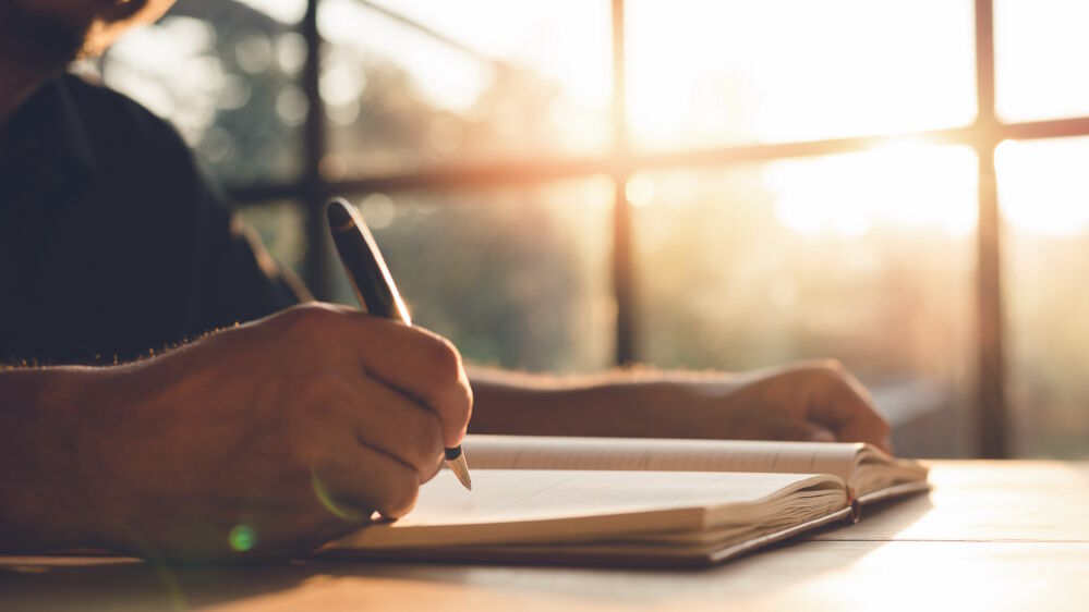 Hand eines Mannes, welcher einen Stift in der Hand hält, um etwas in ein vor ihm liegendes leeres Buch zu schreiben.